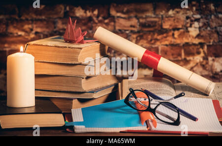 Stapel Bücher mit Origami auf der Oberseite, Kerze, Buch, Notebook, Bleistift und Gläser auf dem Holz braunen Hintergrund gegen alte Mauer Stockfoto