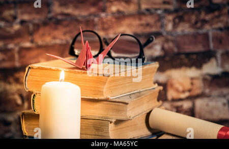 Stapel Bücher mit Origami auf der Oberseite, Kerze, Buch, Notebook, Bleistift und Gläser auf dem Holz braunen Hintergrund gegen alte Mauer Stockfoto