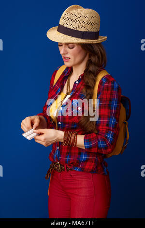Auf der Suche nach inspirierenden Orten. Aktive kranke Frau Wanderer in einen Plaid Shirt nimmt eine Tablette aus der Verpackung auf blauem Hintergrund Stockfoto