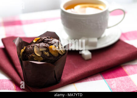 Tee mit Zitrone in einem weißen Schale auf eine Untertasse mit Würfeln von raffinated Zucker und Schokolade Muffin mit Pistazien auf einem roten Serviette Stockfoto