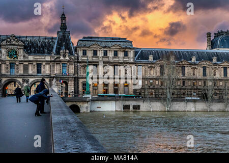 Paar Touristen fotografieren auf der Pont du Carrousel Brücke in Paris, flankiert von Louvre Architektur und einem stimmungsvollen feurige Himmel bei Dämmerung Stockfoto