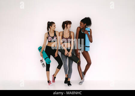 Gruppe von weiblichen Freunde in Sportkleidung zusammen Lächeln, während in der Turnhalle stehen nach Yoga Workout. Frauen, die durch eine Wand mit trainingsmatte. Stockfoto