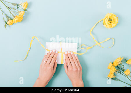 Frau Verpackung ein Geschenk. Blick von oben auf die weibliche Hände mit Ringen das Binden einer Schleife auf die geschenkbox unter gelbe Rosen am trendigen blauen Hintergrund. Flach gesundes Stockfoto