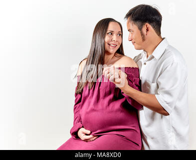 Attraktive junge schwangere Frau das Tragen eines burgandy ärmelloses Kleid sitzen, während ihr Mann suchen und lächelnd. Vater des Babys trägt Stockfoto