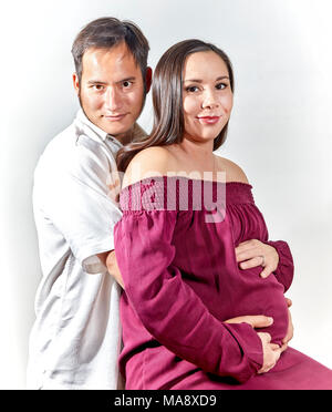 Attraktive junge schwangere Frau das Tragen eines burgandy ärmelloses Kleid mit ihren Händen ruht auf ihrem Bauch beim Sitzen und in die Kamera und Sm suchen Stockfoto