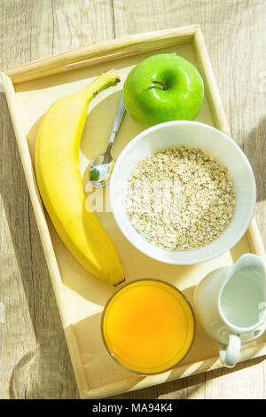 Holz- Fach mit gesunden Zutaten für das Frühstück auf dem Tisch. Hafer in der Schüssel Mutter Milch in Krug Orangensaft Banane Green Apple. Helle Morgensonne Str Stockfoto