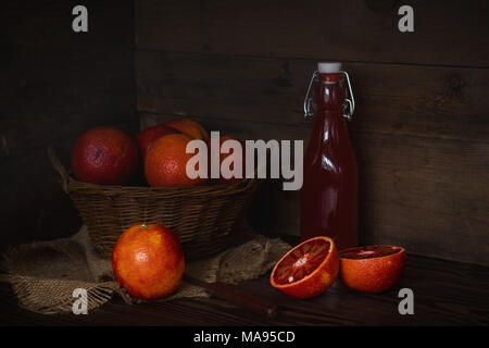 Blut orange Frucht in einem Weidenkorb und Flasche Saft auf dunklen Holztisch. Dunklen rustikalen Stil. Stockfoto