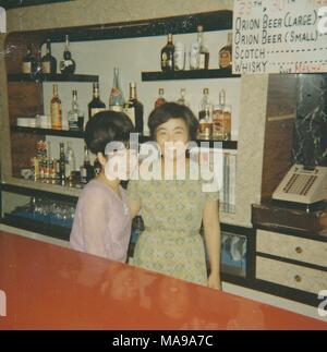 Farbfoto, zeigt zwei Frauen, hinter dem Tresen einer Bar, mit Flaschen Alkohol und eine Registrierkasse im Hintergrund, eine Werbung für Orion Bier sichtbar, Okinawa, Japan, 1968. () Stockfoto