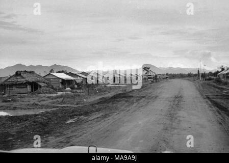 Schwarz-weiß Foto zeigt eine unbefestigte Straße mit einer Reihe von kleinen Gebäuden, Häusern, eines mit Reetdach, auf beiden Seiten der Straße, die in Vietnam während des Vietnam Krieges fotografiert (1955-1975), 1968. () Stockfoto