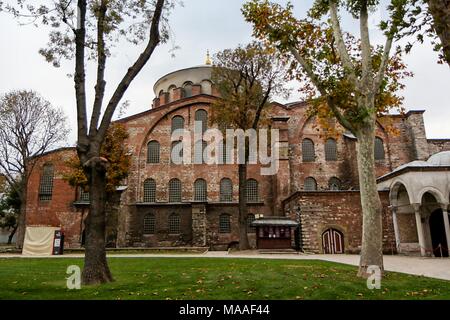 Außenansicht, Gras und zwei Bäume im Vordergrund und im Hintergrund, die Hälfte der im osmanischen Stil, gewölbt und gewölbten Eingang Vorhalle (rechts) und der südlichen Fassade aus Stein und Backstein gebaut Aya Irini (Hagia Irene), eine ehemalige Griechische Orthodoxe Kirche, jetzt als Museum und Konzertsaal, im äußeren Hof des Topkapi-Palastes in Istanbul, Türkei, 16. November 2017 verwendet. () Stockfoto