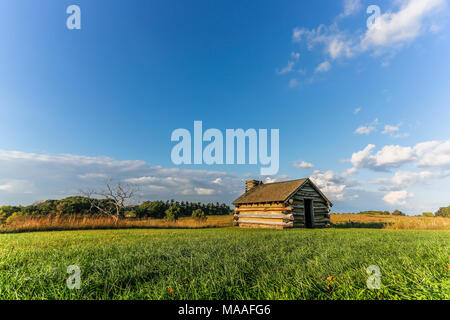 Eine einsame Blockhütte sitzt allein in der Mitte einer großen hellen, offenen Landschaft von Himmel und Felder bis an den Horizont an einem sommerlichen Nachmittag; mit Cop Stockfoto