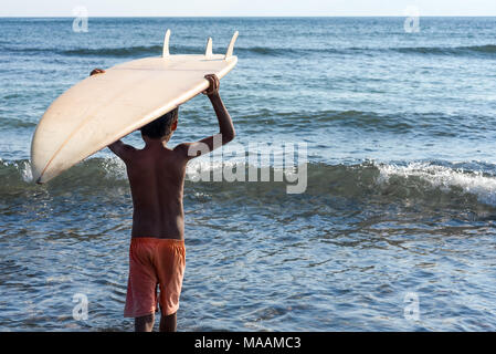 Die jungen lokalen Indonesischen Junge in verschlissenen Shorts trägt seinen gespendet Surf Board auf seinem Kopf in einem ruhigen Meer. Stockfoto