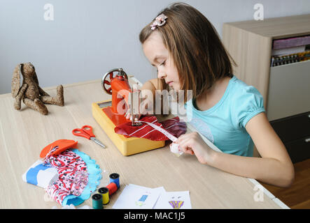 Das Mädchen ist das Nähen eines Kleides für eine Puppe auf einer Nähmaschine. Sie mit einem Maßband, das Produkt zu messen. Stockfoto