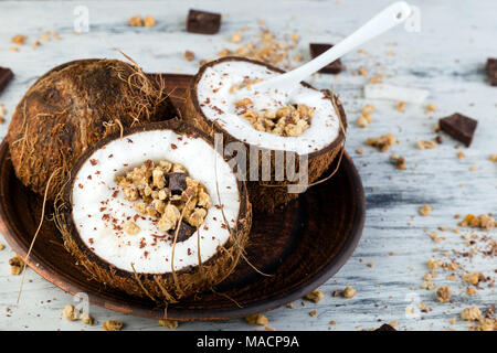 Gesundes Frühstück in Coconut Bug auf weißem Hintergrund. Joghurt in Kokosnuss Schale mit Kokosraspeln, Schokolade und Müsli. Ansicht von oben, flach, Overhead Stockfoto