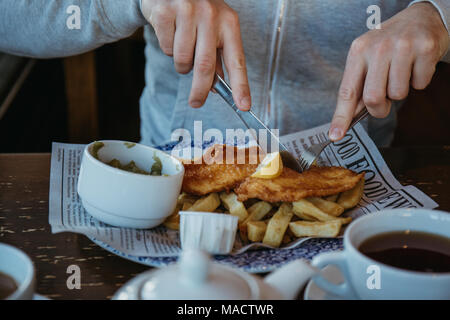 Mann am Tisch sitzen, essen Fisch und Chips, traditionelle englische Gerichte, Kaffee und Saucen in der Nähe. Stockfoto