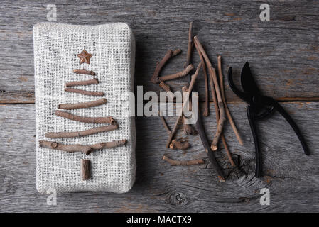 Overhead shot eines Sackleinen gewickelt Weihnachtsgeschenk mit einem Zweig, Baum und Stern. Verschiedene Zweige und Baum-, Reb-, Gartenscheren sind auch auf der rustikalen, mit Holz Tisch. Stockfoto