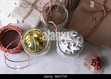 Weihnachtsgeschenk Verpackung liefert. Hohe Aufnahme von drei Maurergläser mit Geschenkanhänger, Holz Sterne, und Schlittenglöckchen gegen eine weiße Wand. Stockfoto