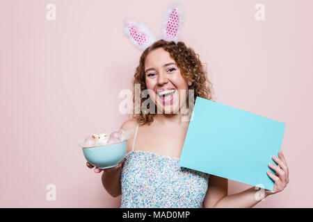 Schöne Frau halten Ostern Dekorationen und ein leeres Plakat mit Hasenohren auf rosa Hintergrund mit kopieren. Stockfoto