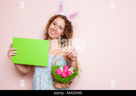 Schöne Frau halten Ostern Dekorationen und ein leeres Plakat mit Hasenohren auf rosa Hintergrund mit kopieren. Stockfoto