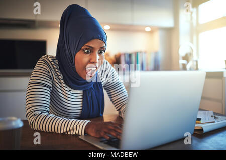 Junge arabische weibliche Unternehmer trägt ein Kopftuch sitzen an ihrem Küchentisch mit einem Laptop und überrascht, während die von zu Hause aus arbeiten Stockfoto