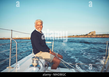 Lächelnd reifer Mann allein sitzen auf dem Deck des Schiffes segeln entlang der Küste an einem sonnigen Nachmittag Stockfoto