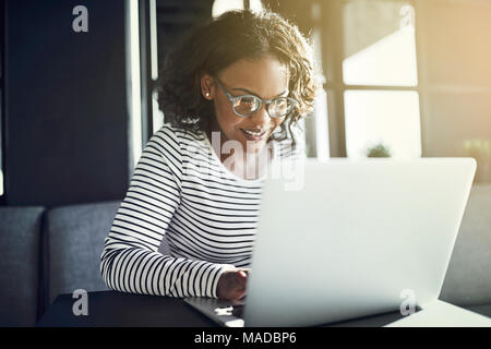 Lächelnden jungen afrikanischen Frau Brille trägt allein sitzen an einem Tisch mit einem Laptop Stockfoto
