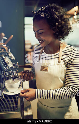Lächelnden jungen afrikanischen barista Gießen eine Tasse frischen Kaffee und eine Kaffeemaschine in einem angesagten Café stehend Stockfoto