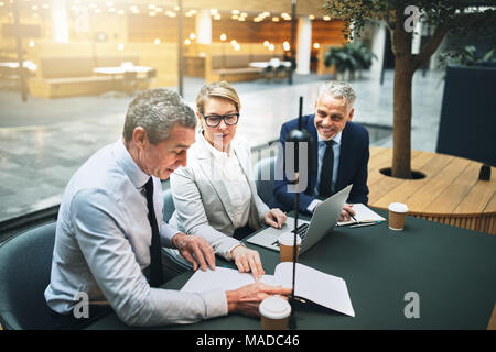 Drei ältere Führungskräfte diskutieren Schreibarbeit und über einen Laptop während einer Sitzung zusammen, die in der Lobby eines Bürogebäudes Stockfoto