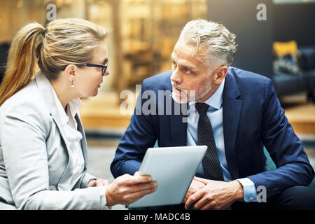 Reifen Geschäftsmann und Geschäftsfrau, sitzen auf Stühlen in einem Büro Lobby reden und mit Hilfe eines digitalen Tablet konzentriert Stockfoto
