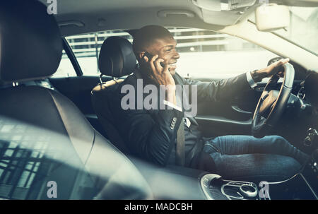 Lächelnd afrikanischen Geschäftsmann trägt einen Blazer Gespräche am Mobiltelefon, während sein Auto durch die Stadt fahren während seiner Morgen pendeln zur Arbeit Stockfoto