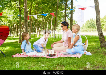 Drei Mädchen und junge Frau mit Picknick auf dem grünen Rasen in öffentlichen Park nicht weit von Spielplatz Stockfoto