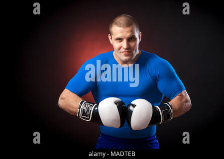 Eine starke Dunkelhaarige sportman Boxer in einem blauen Sportbekleidung rush Guard und in Schwarz und Weiß Boxhandschuhe, gegen einen roten Lampen auf einem schwarzen Stockfoto