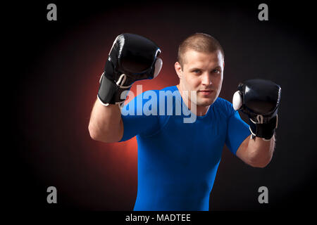 Eine starke Dunkelhaarige sportman Boxer in einem blauen Sportbekleidung rush Guard und in Schwarz und Weiß Boxhandschuhe Boxen gegen einen roten ein Licht auf einem schwarzen i Stockfoto