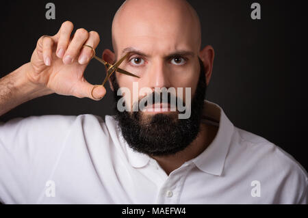 Portrait des Kahlen bärtigen Mann mit Schere auf sein Auge über dunklen Hintergrund Stockfoto