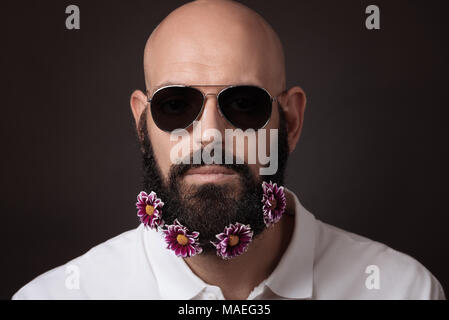 Portrait von unrasierten Mann mit Bart und Schnurrbart mit chrysantheme Blumen und eine dunkle Sonnenbrille auf dunklem Hintergrund Stockfoto