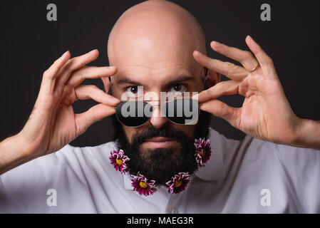 Portrait des Kahlen unrasierten Mann mit chrysantheme Blumen auf den Bart und mit Sonnenbrille auf dunklem Hintergrund Stockfoto