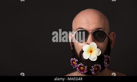 Portrait des Kahlen unrasierten Mann mit Bart und Schnurrbart mit Chrysanthemen und Magnolia Blumen und Sonnenbrille auf dunklem Hintergrund mit Copyspace Stockfoto
