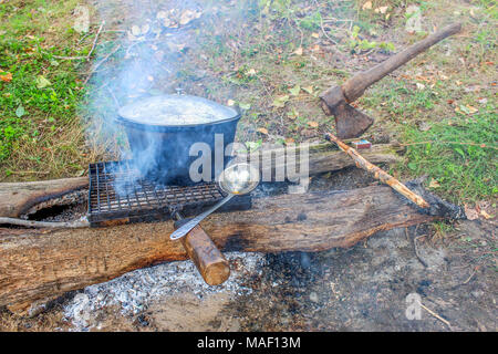 Aluminium Topf ist über dem Kamin auf den Kohlen beheizt, stehend auf dem Grill für ein Barbecue auf zwei Protokolle und eine Axt stecken in einer Protokolldatei Stockfoto