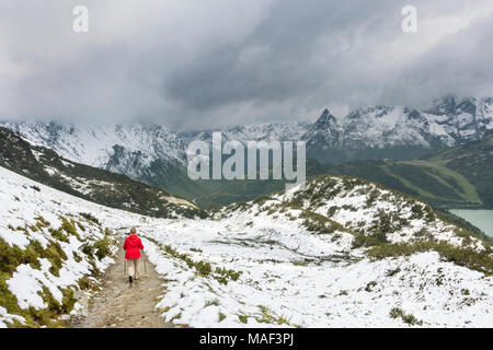 Eine weibliche ältere Wanderer trägt einen roten Mantel wandern durch Schnee im Sommer auf dem Weg zum Zeinisjoch im Paznauntal in der Nähe von Galtür in Österreich. Die Stockfoto