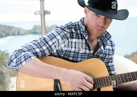Schöne Country und Western Cowboy Sänger mit Hut und Gitarre spielen auf hotel Balkon mit Blick aufs Meer im Hintergrund Stockfoto
