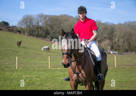 Stattliche männliche Pferd Reiter zu Pferd im grünen Feld mit Pferden und Landhaus aus Stein im Hintergrund. Mit schwarzen Helm, Stiefel weiß Hose Stockfoto