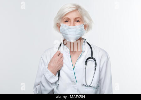 Im Alter von Frau Doktor tragen Mundschutz Studio auf weißem Hintergrund holding Stethoskop Lächeln mit den Augen isoliert Stockfoto
