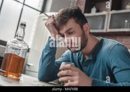 Junger Mann rauchen Suchtprobleme bei Tisch holding Zigarette sitzen Suche im Whiskey Glas umgekippt Nahaufnahme Stockfoto