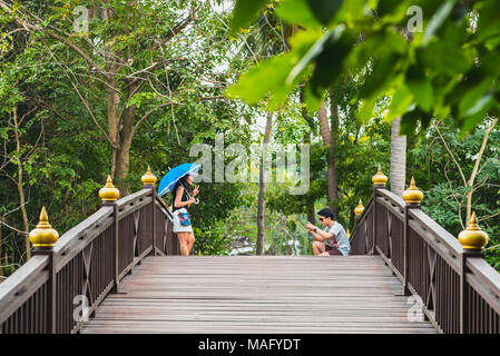 Junge asiatischer Mann Fotografien eine lächelnde junge asiatische Frau mit blauen Dach auf hölzernen Brücke im Park im Bang Krachao (Bang Kachao), Bangkok, Thailand. Stockfoto