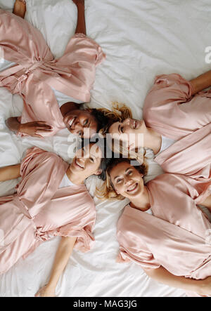 Blick von oben auf die Braut mit Brautjungfern liegend auf Bett und lächelt. Gruppe von Frauen mit Schlafrock auf Bett zu Hause liegen. Stockfoto