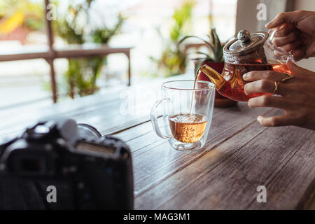 Nahaufnahme einer Frau gießen Kaffee in eine Tasse aus einer Teekanne aus Glas. Frau die Zubereitung von Tee, während eine professionelle Kamera und einem Blumentopf auf dem Tisch platziert werden Stockfoto