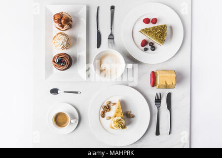 Blick von oben auf die Anordnung der Stücke von verschiedenen Kuchen auf Tellern, Tassen Kaffee und Muffins isoliert auf weißem Stockfoto