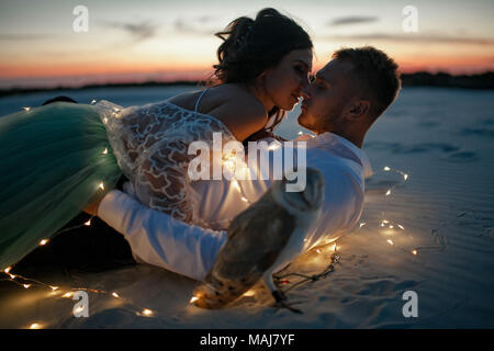 Braut und Bräutigam liegen auf Sand in der Wüste und Kiss neben Girlande aus leuchtenden Glühbirnen und owl gegen die untergehende Sonne. Nahaufnahme. Stockfoto