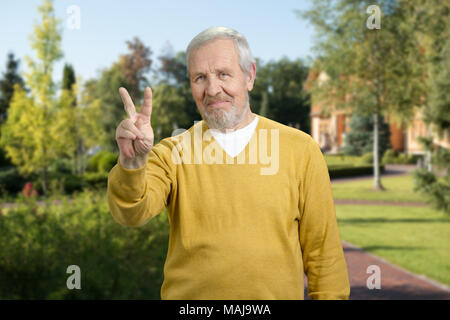 Alter Mann mit Sieg Geste. Senior zeigt zwei Finger outdoor. Bäume im Sommer Hintergrund. Stockfoto