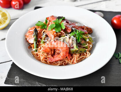 Spaghetti Frutti di Mare oder Pasta mit Garnelen und grünem Spargel und Petersilie garnieren. Weiße Platte auf einem Holztisch, gesunde Ernährung Szene. Stockfoto